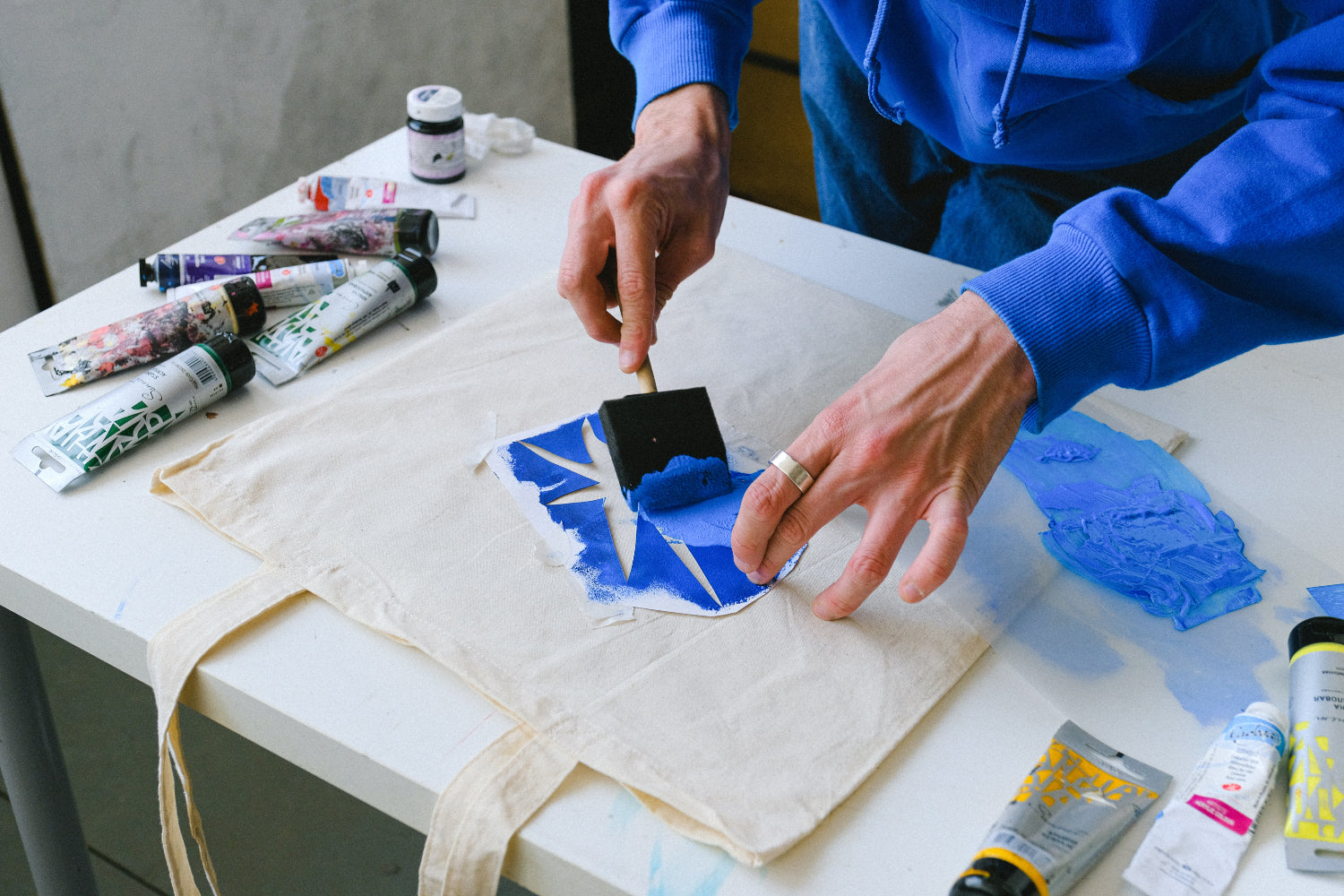 A person makes a merch tote bag using a stencil and blue paint