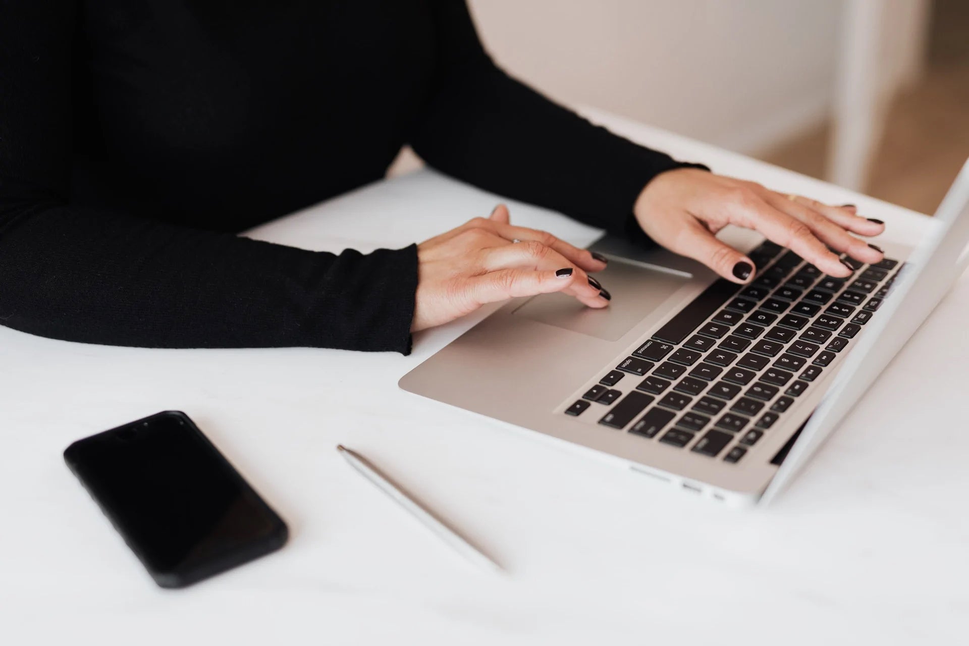 A person types on a laptop. A phone and pen sit on the desk beside it