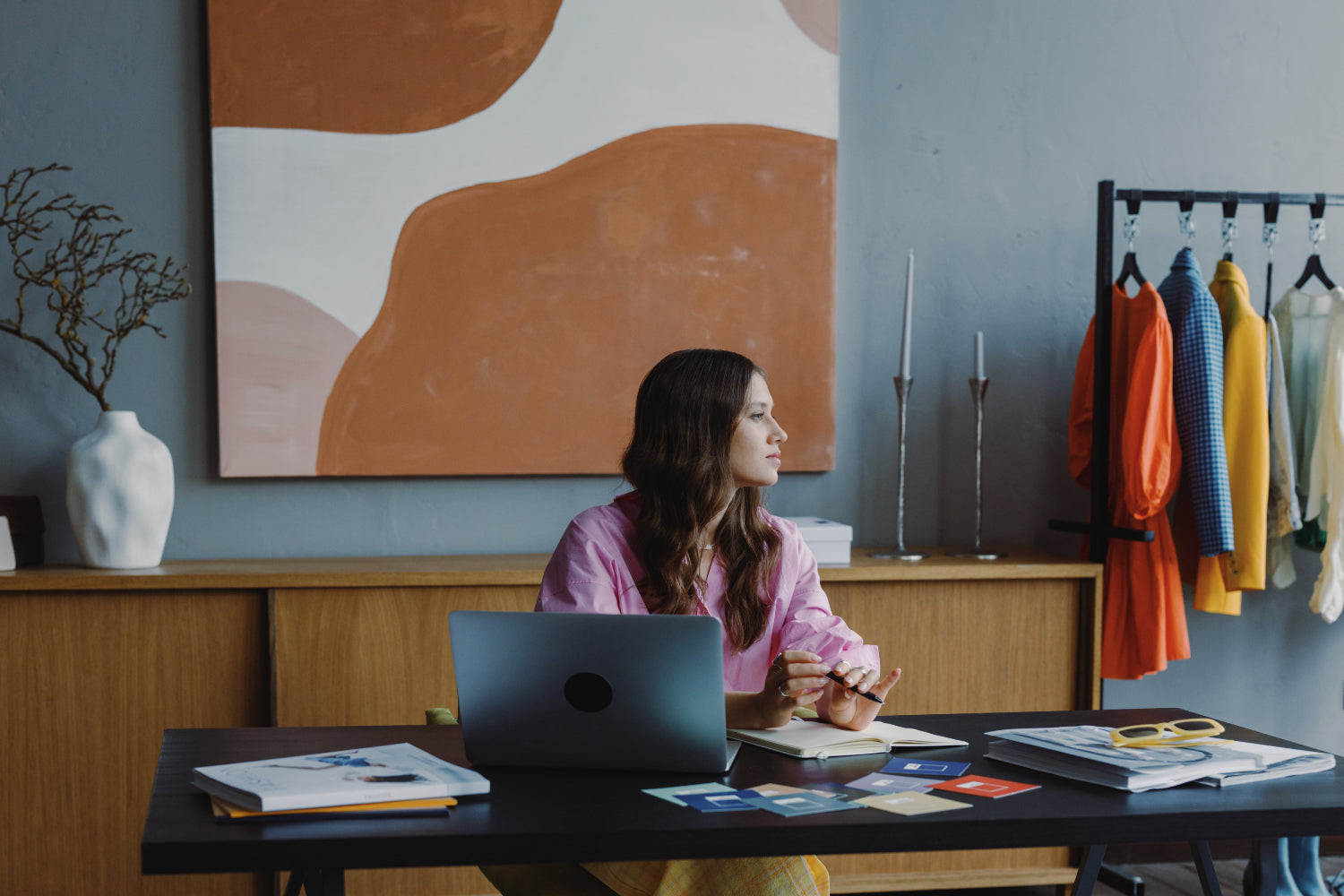 A woman sits at a desk thinking in front of a laptop. A garment rack sits to the right