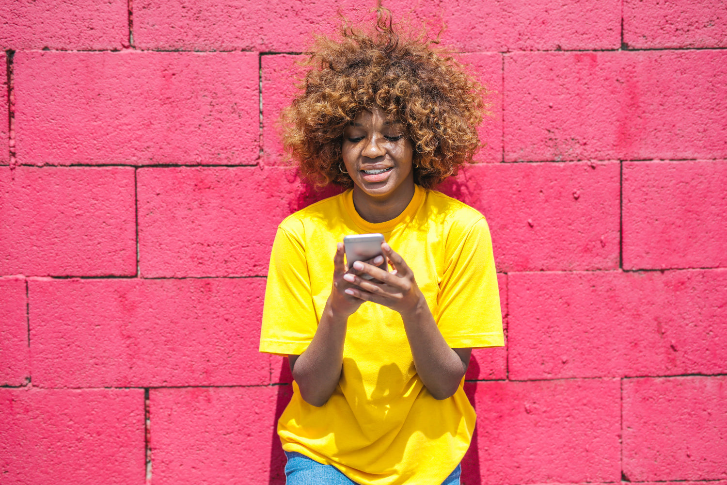 Woman poses against a pink brink wall using a mobile phone