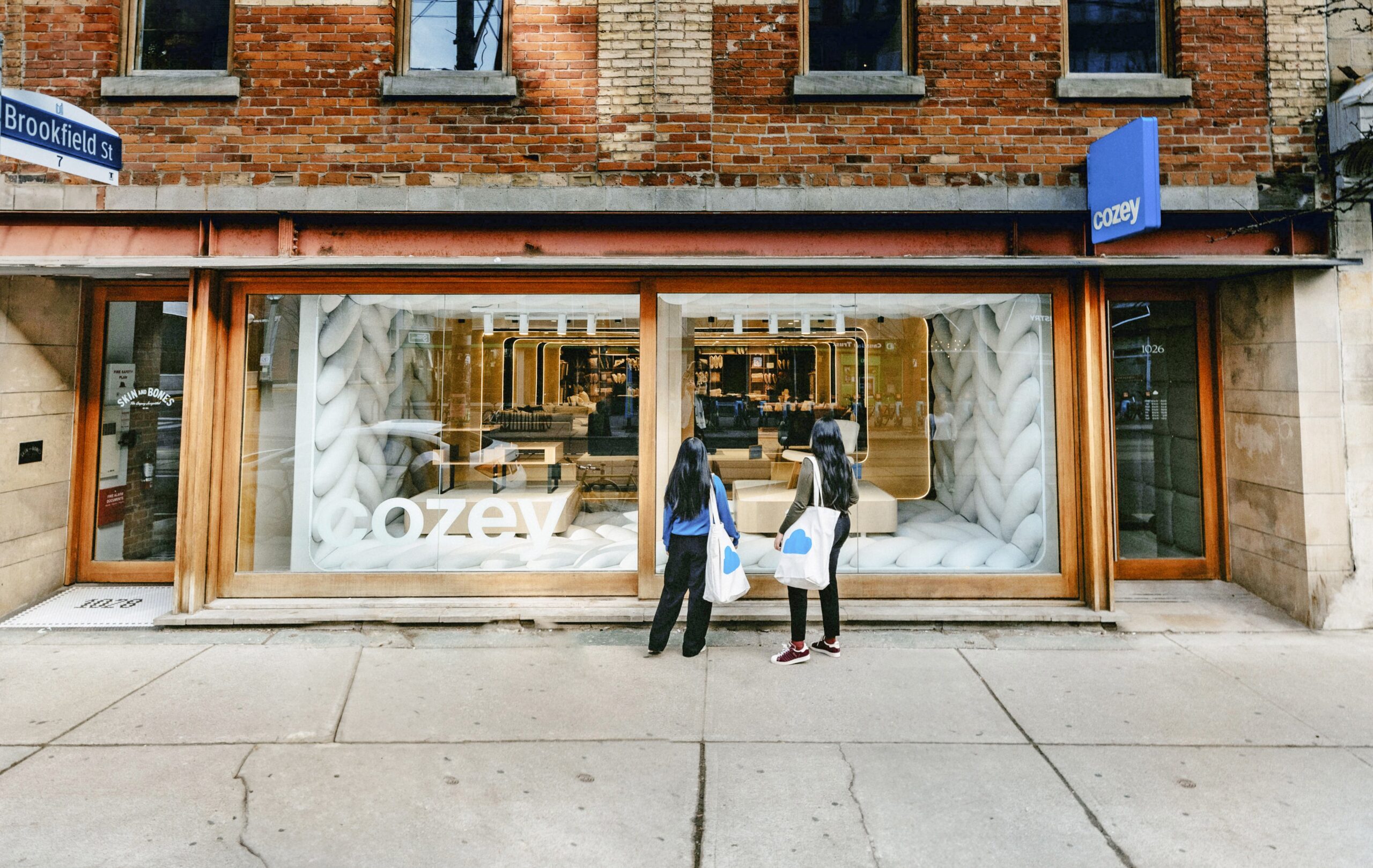 View of Cozey store from the sidewalk, two shoppers are outside the big picture windows wearing tote bags and looking in.