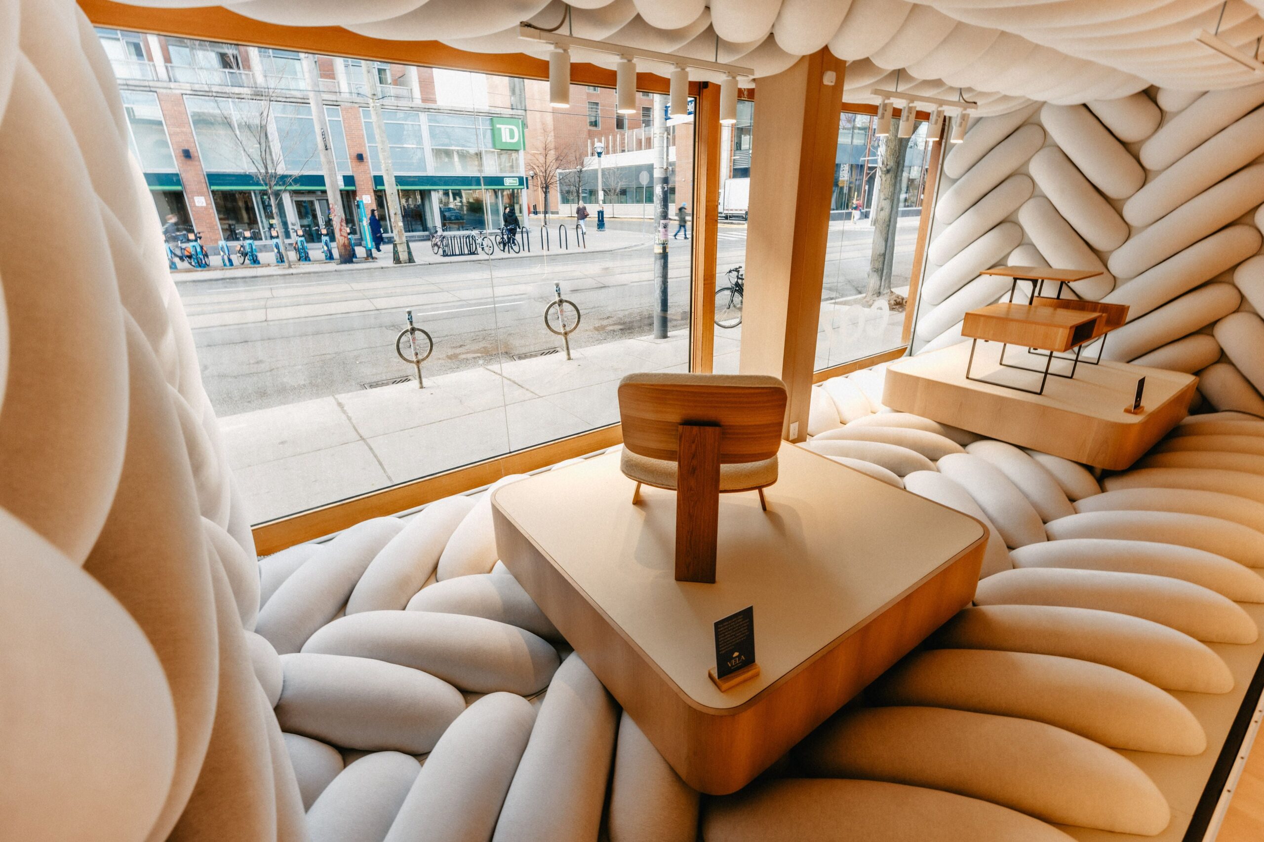 View from within the Cozey store retail window out to the sidewalk with cushions and a platform for a wooden chair and a coffee table.