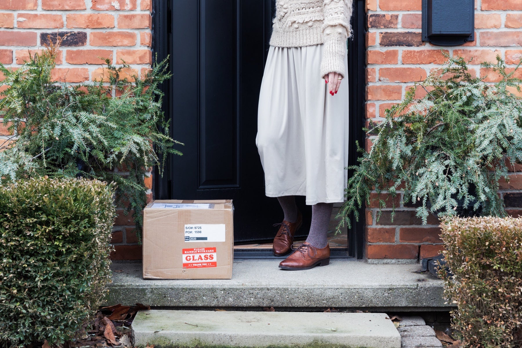 Woman stands at a front door flanked by bushes. A package waits for her on the stoop