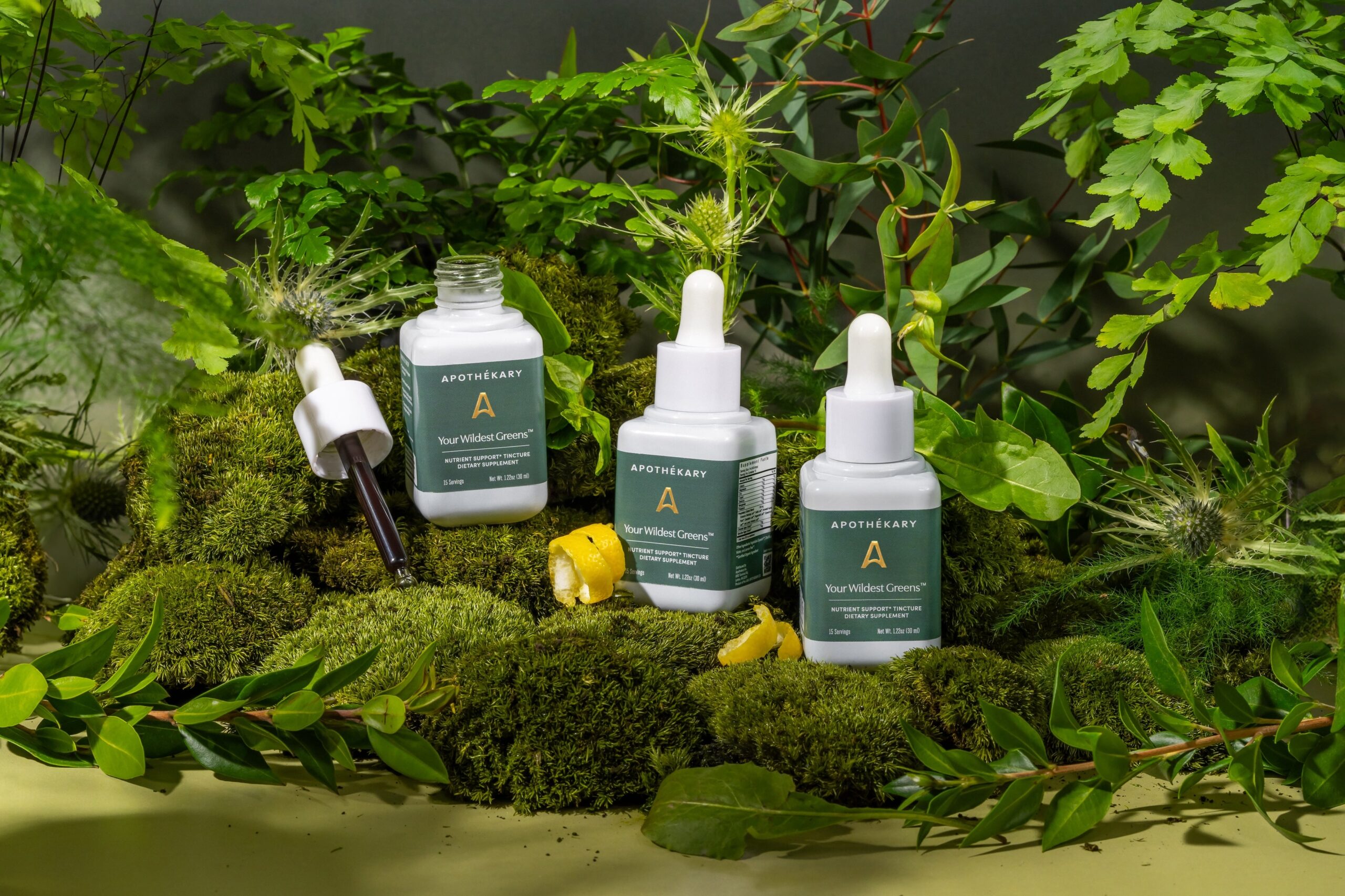 three of apothékary’s your wildest dreams tincture sitting in a sea of green plants with some lemon peels in the forefront.