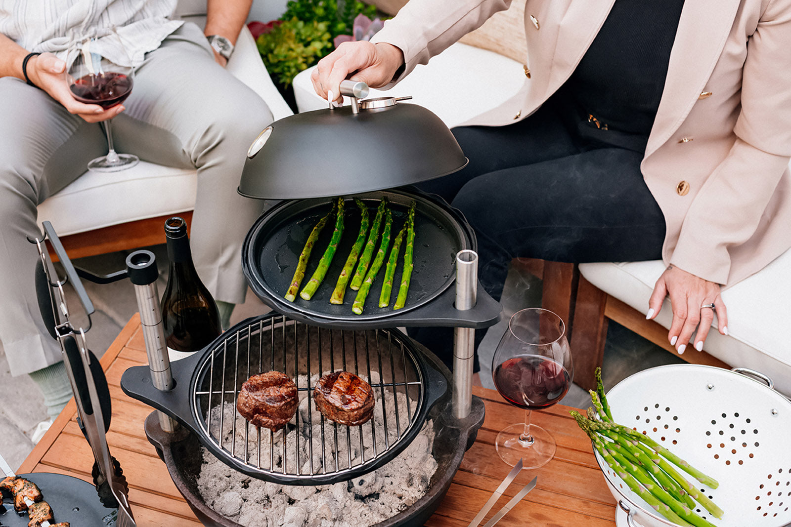 Two people sitting around a Bola Grill with burgers and asparagus on different grill levels.