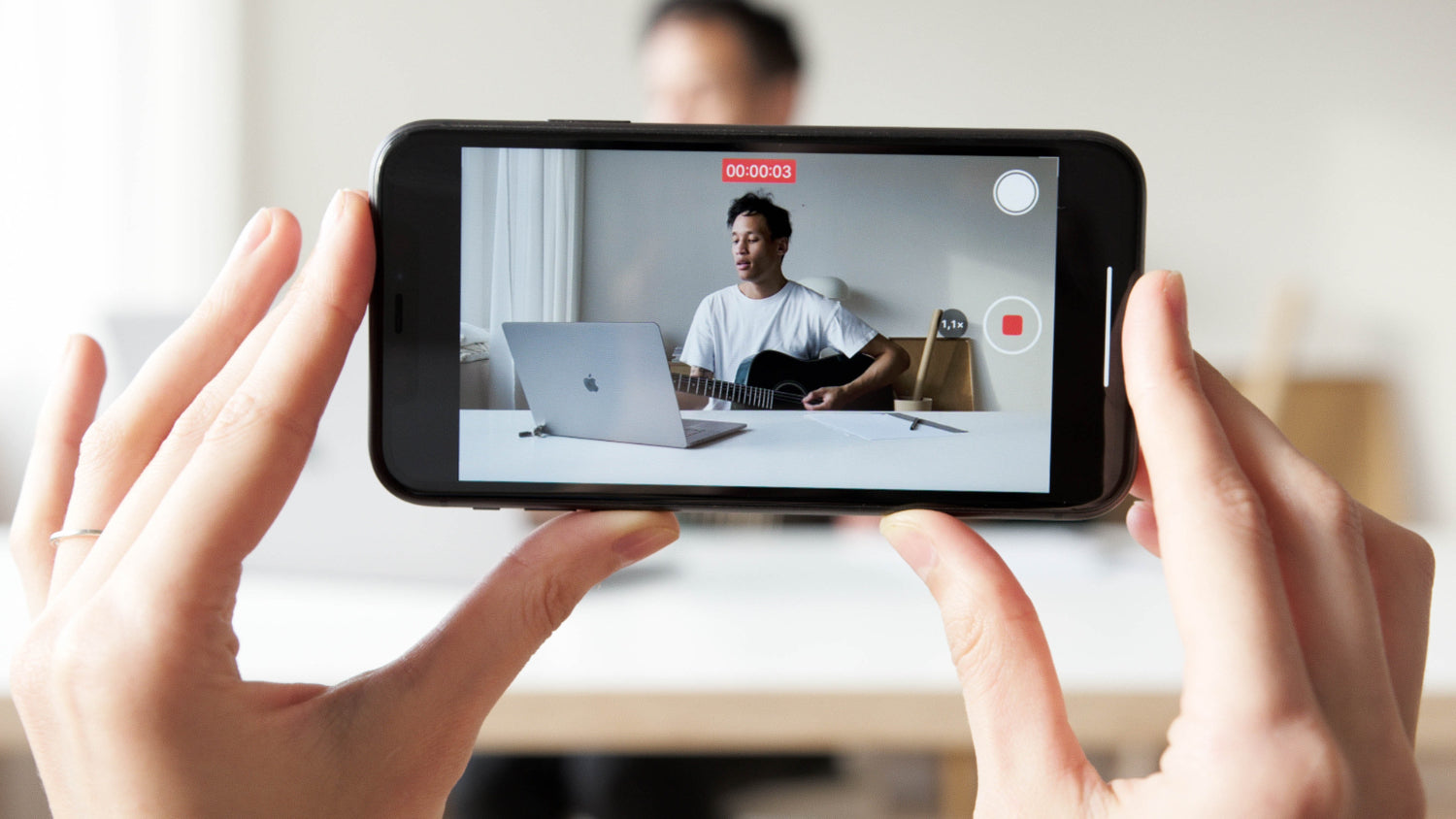 Two hands holding an IPhone horizontal with camera view set to record a guitarist at a laptop.