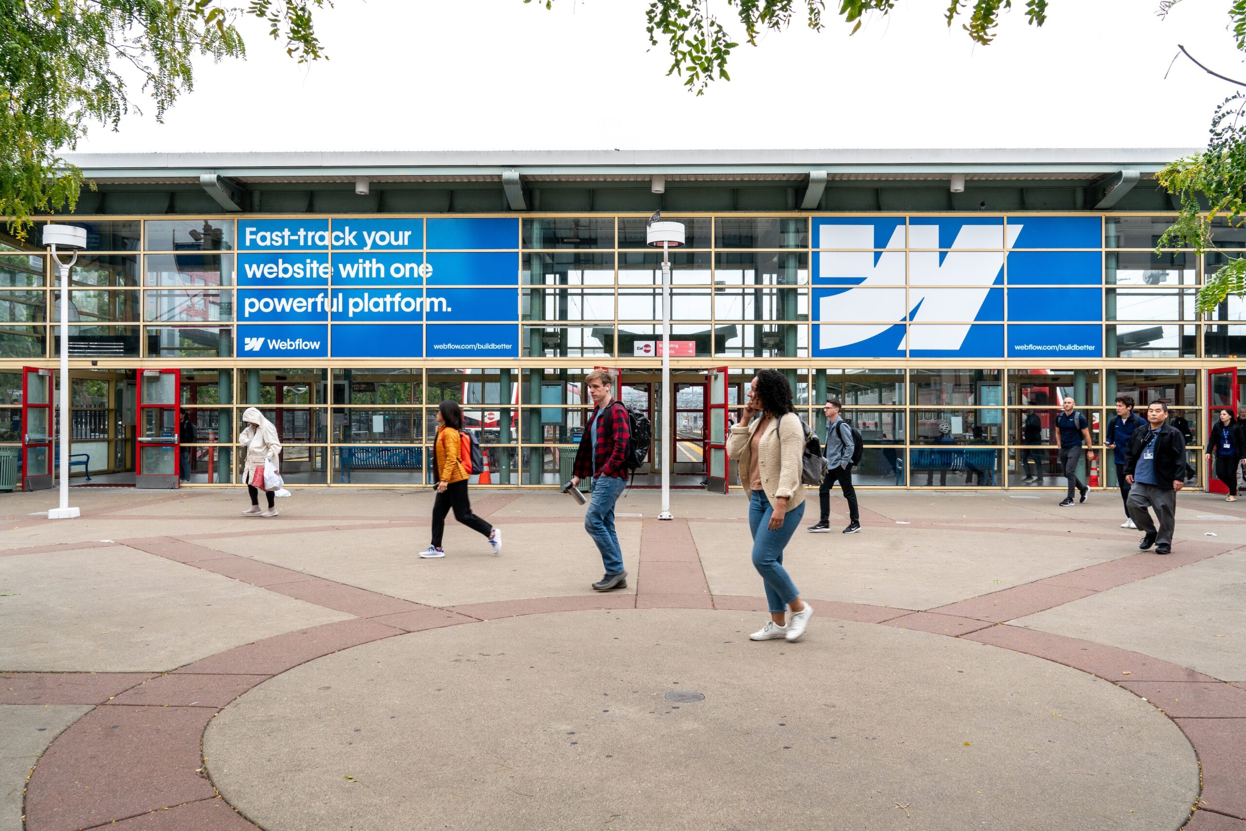 A photo of a train station in San Francisco with people walking out of it. The front windows of the train station display large Webflow ads.