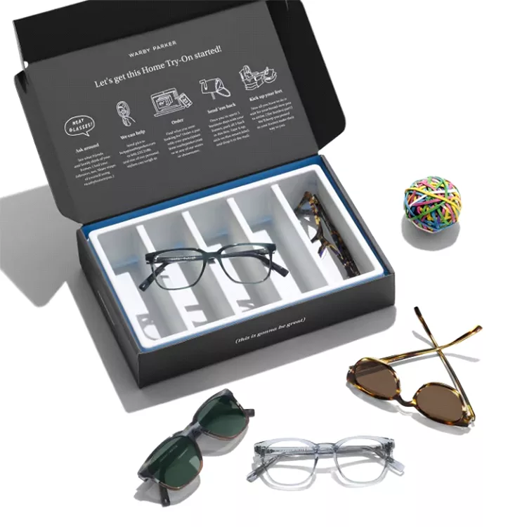 A black box with compartments holds eyeglasses and includes instructions for a home try-on kit. Four pairs of glasses and a rubber band ball are placed around the box.