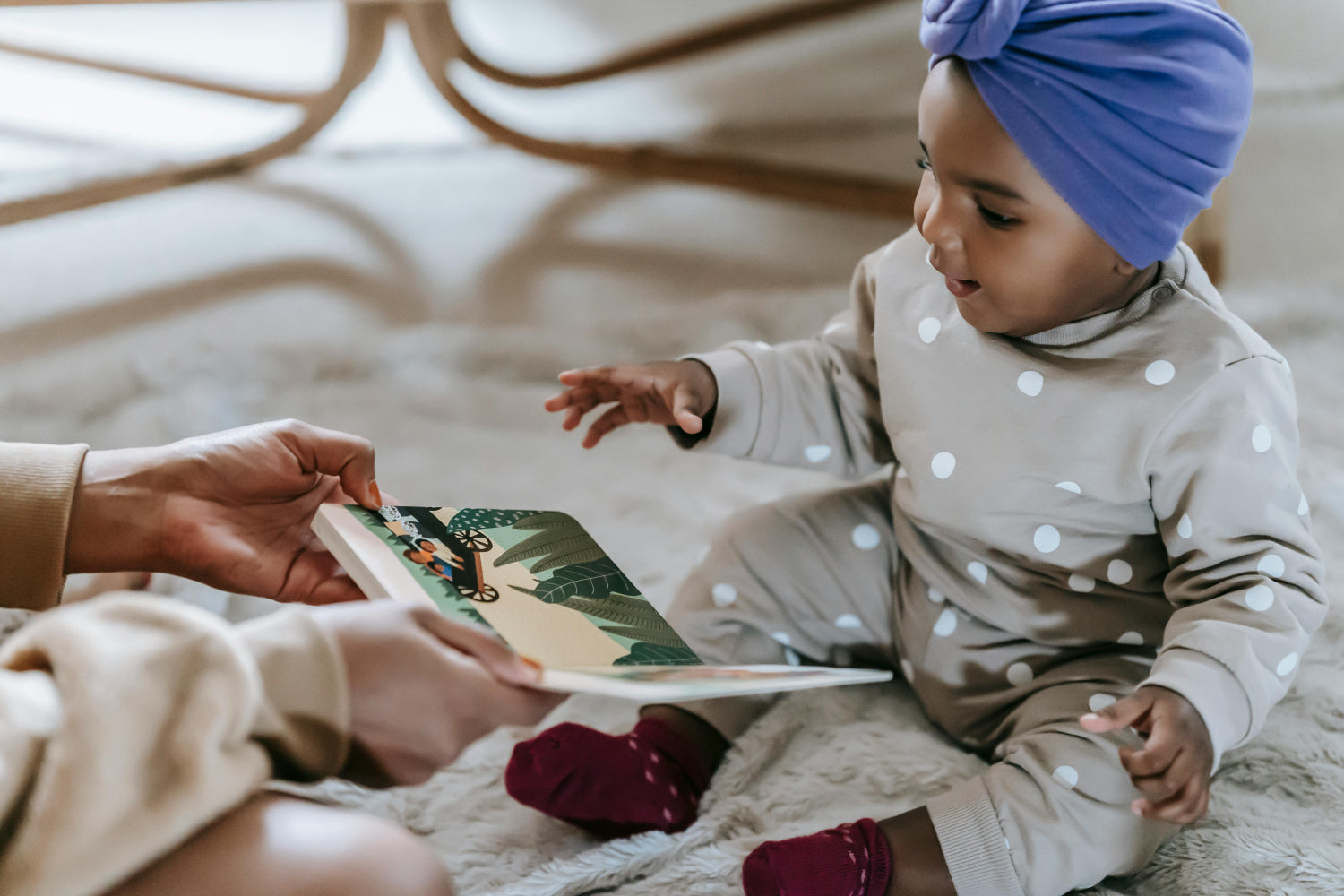 Baby in a polka dot outfit, sitting on a blanket and reaching for a book.