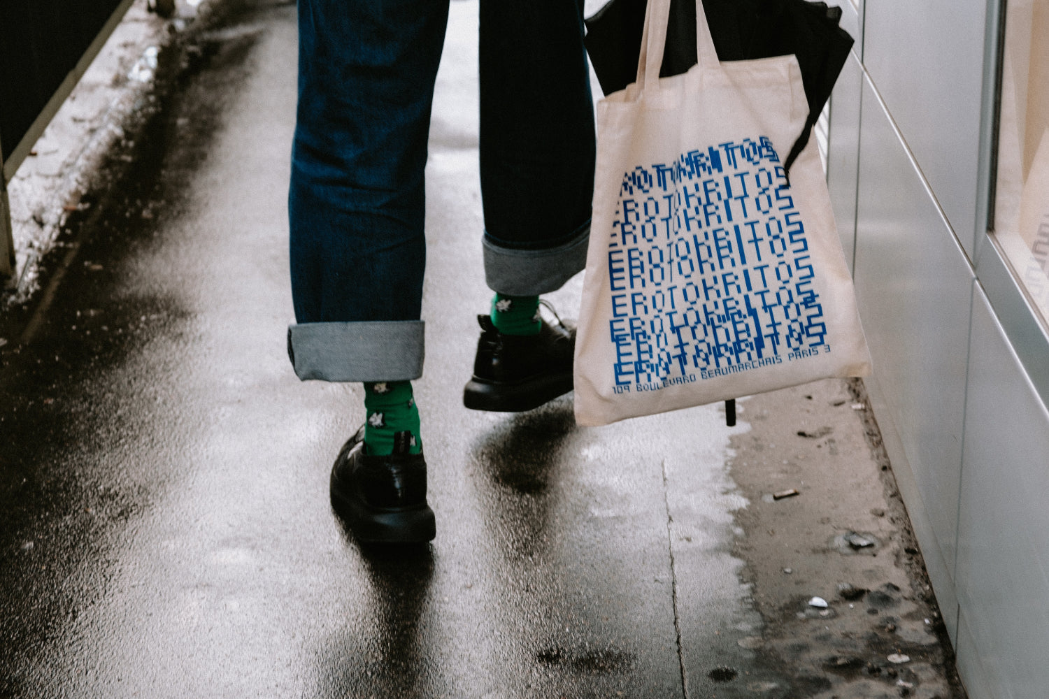 Person walking with a tote bag and green socks, visible from the waist down on a wet sidewalk.