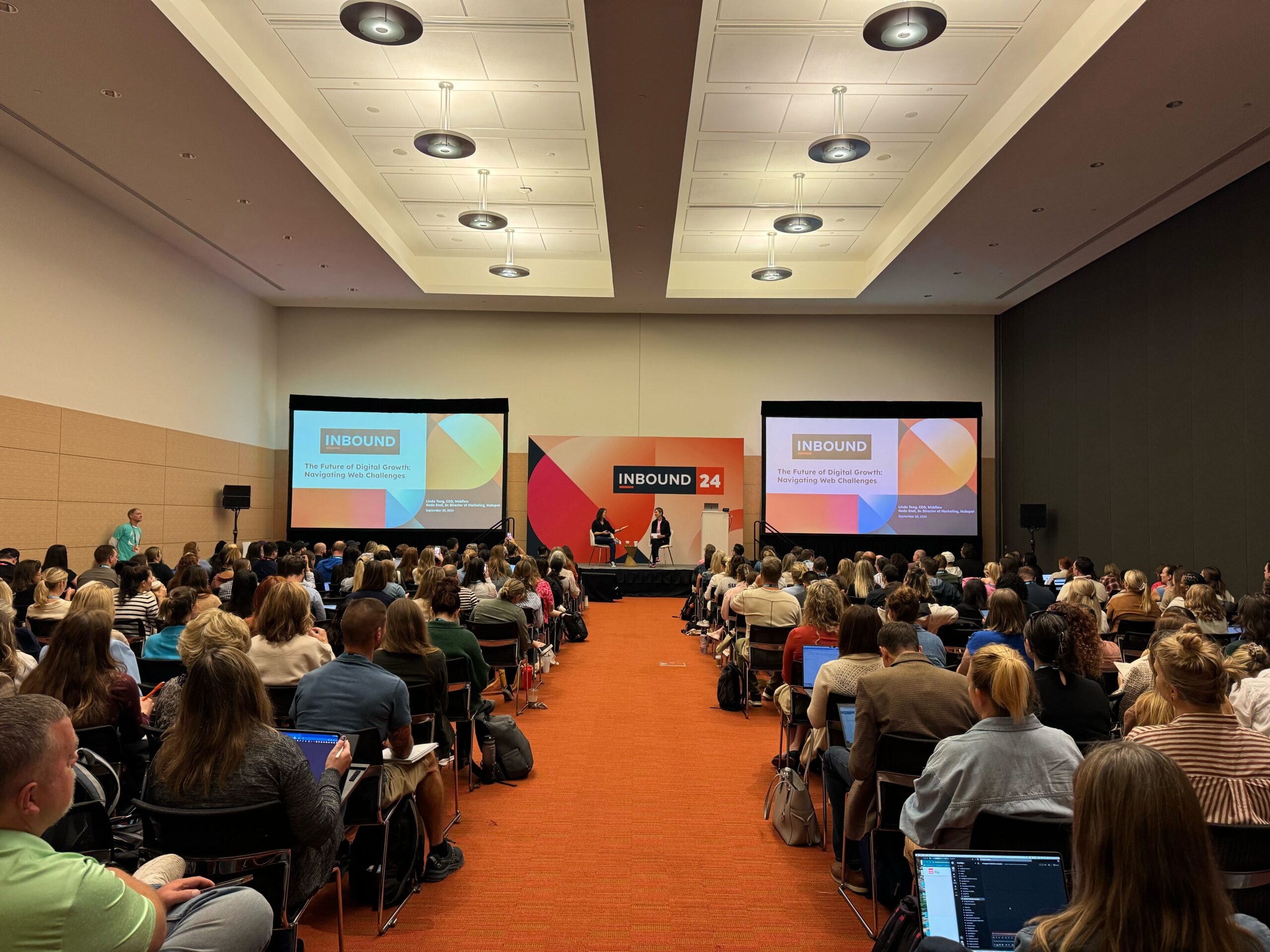 A presentation hall with two women presenting on stage to a room full of seated attendees.