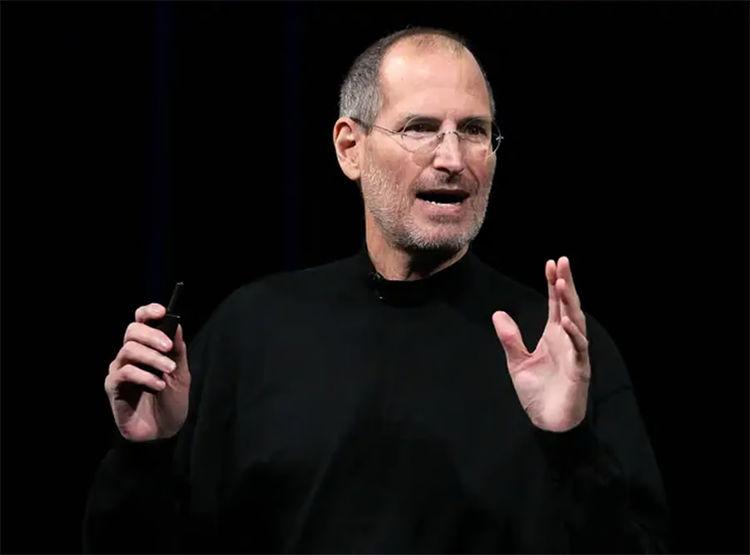 A man wearing glasses and a black turtleneck speaks while gesturing with one hand and holding a small object in the other.