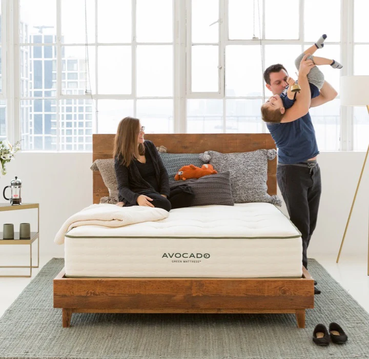 A family relaxes on a bed, with a child happily playing nearby, showcasing a warm family moment.