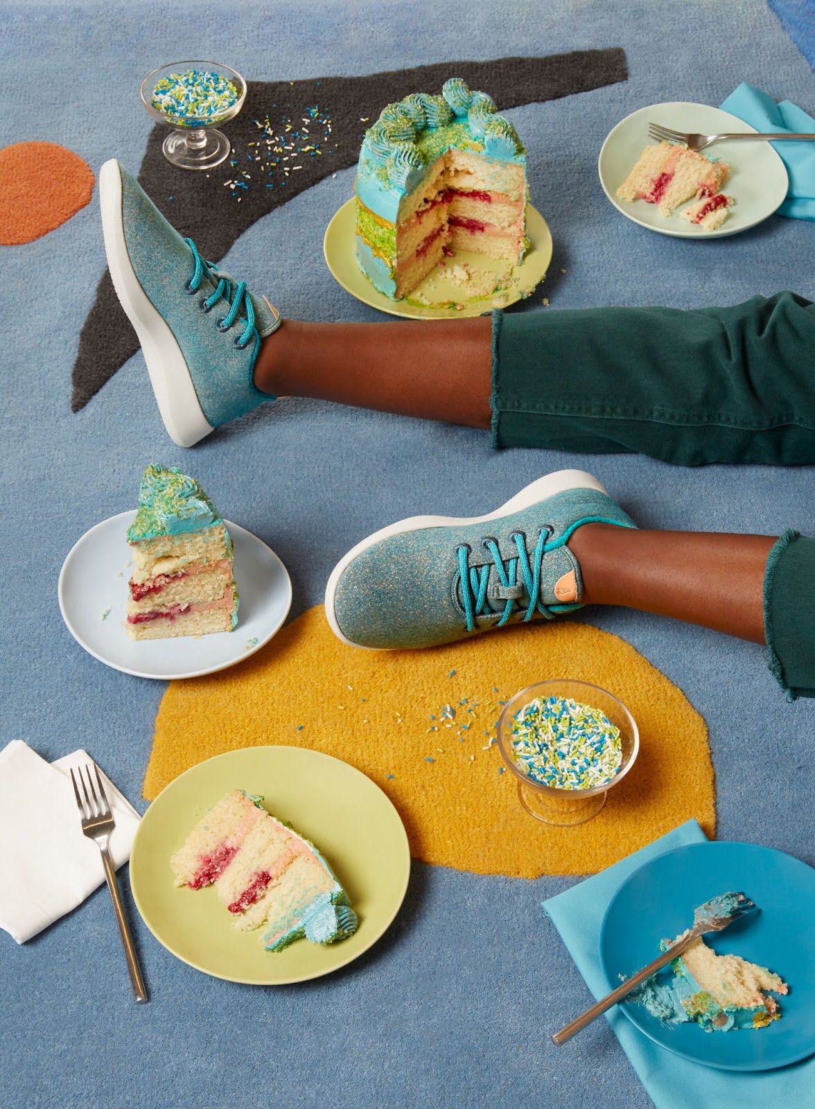 The lower half of a sitting person wearing blue shoes surrounded by cake slices with blue frosting.