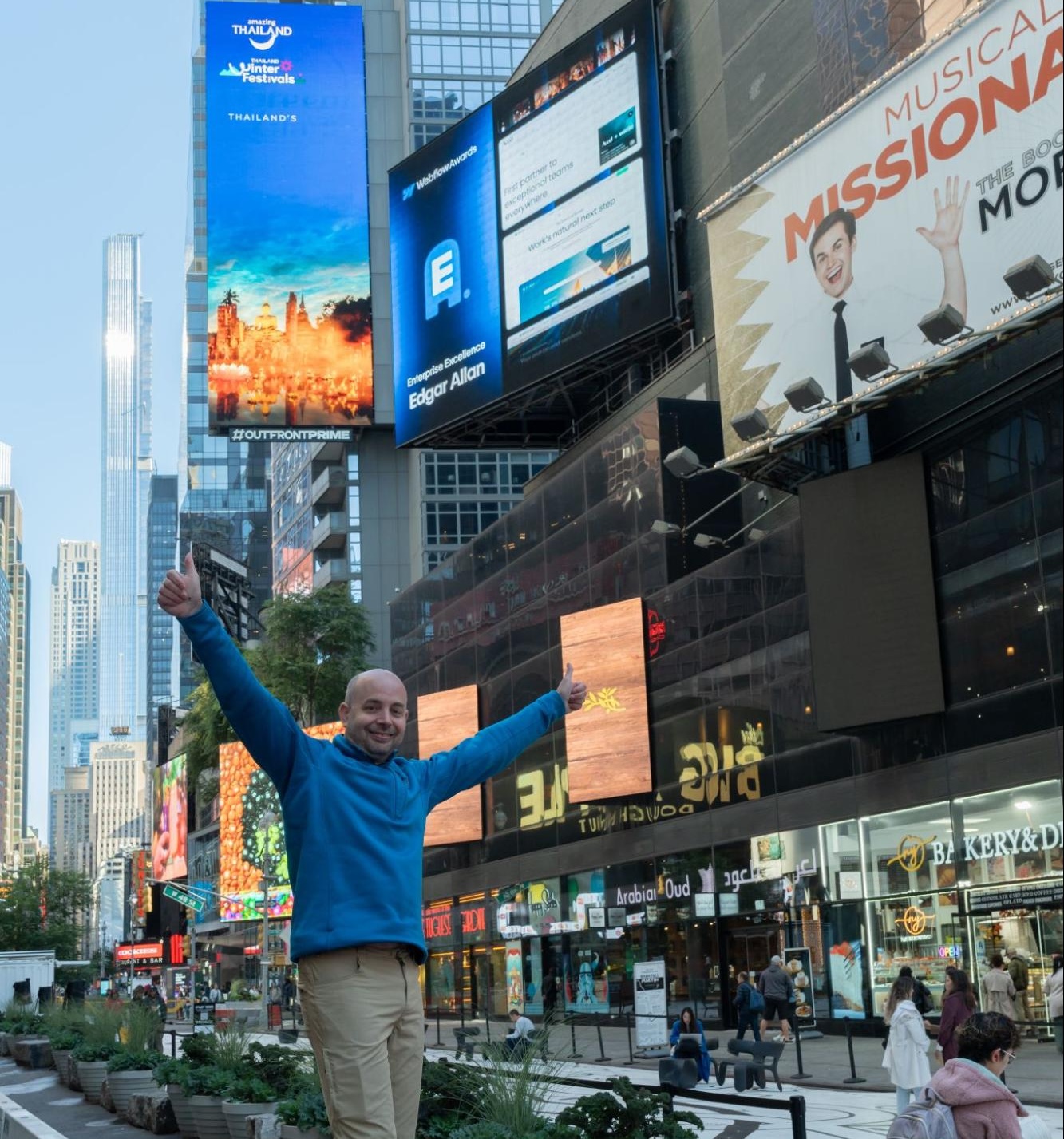 man smiling with his arms raised in front of his company's billboard in times square