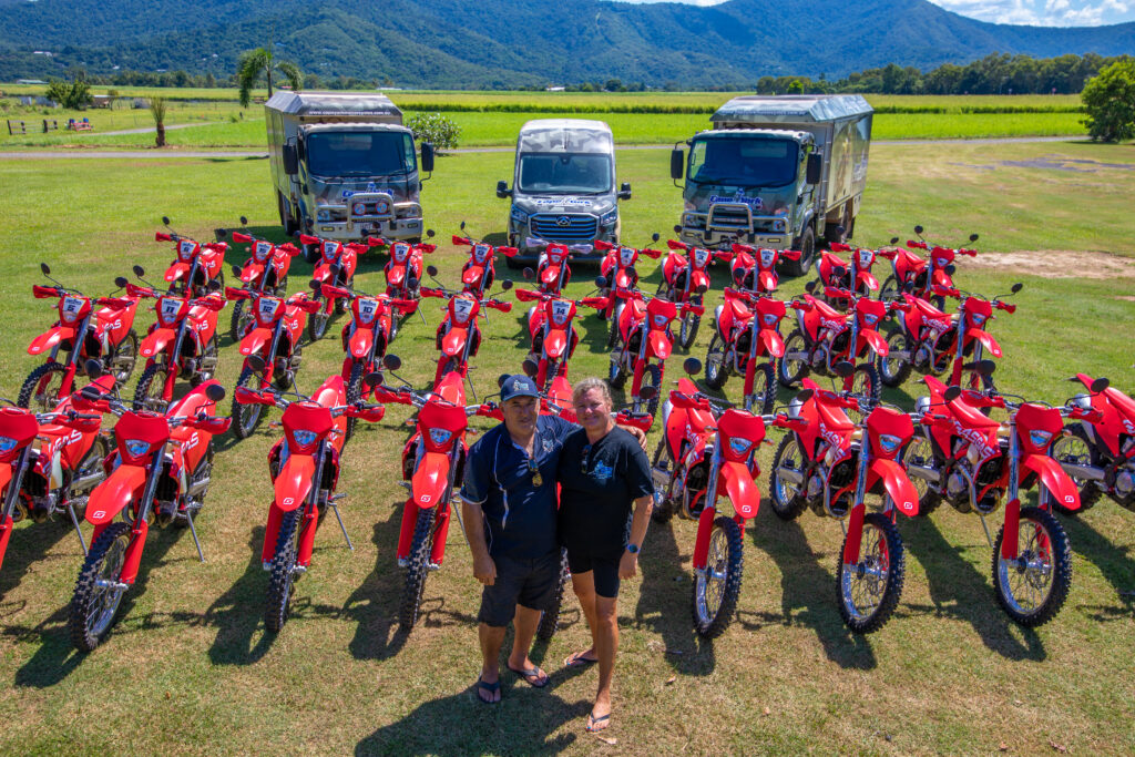 two people standing in front a fleet of motorbikes with three trucks in the background