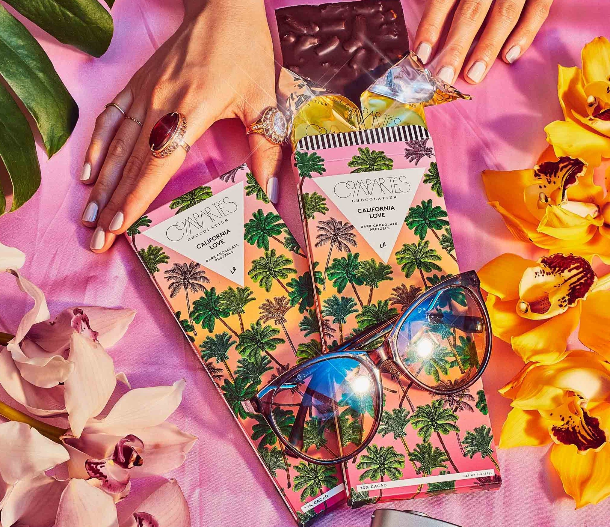 two Compartés California love pretzel chocolate bars, with a model’s hands holding one with sunglasses and plants surrounding them.