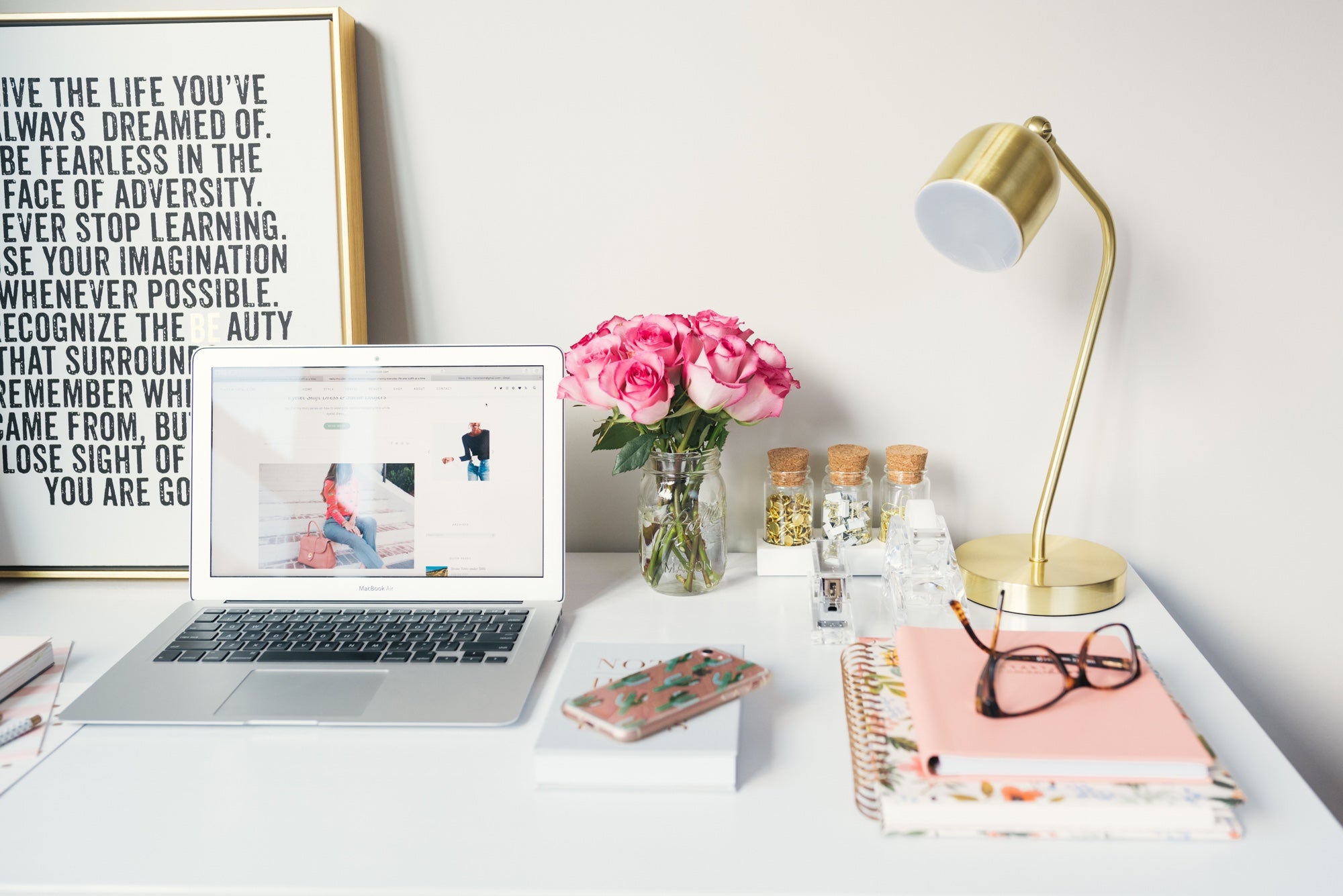 A desk featuring a laptop, lamp, art, and a vase of flowers