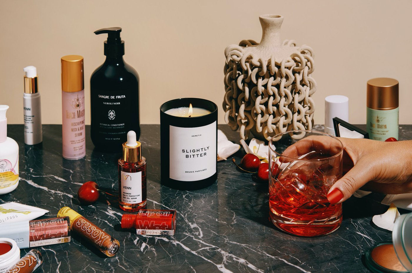 An array of cosmetics products on a table from India Rose. A hand holding a drink enters the frame