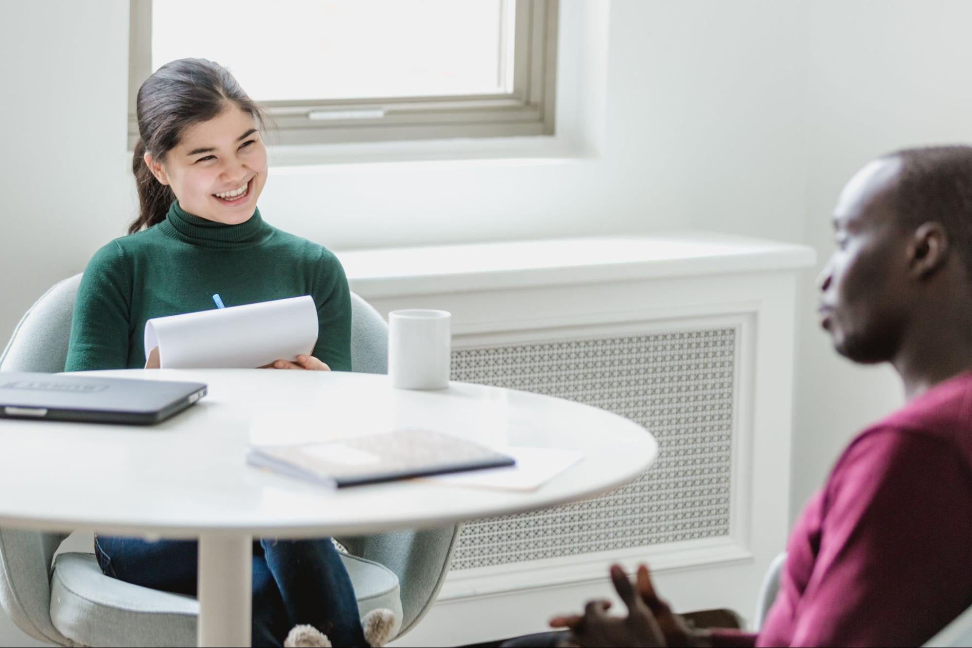 A woman smiles while holding a clipboard and listens to a man sitting across from her.