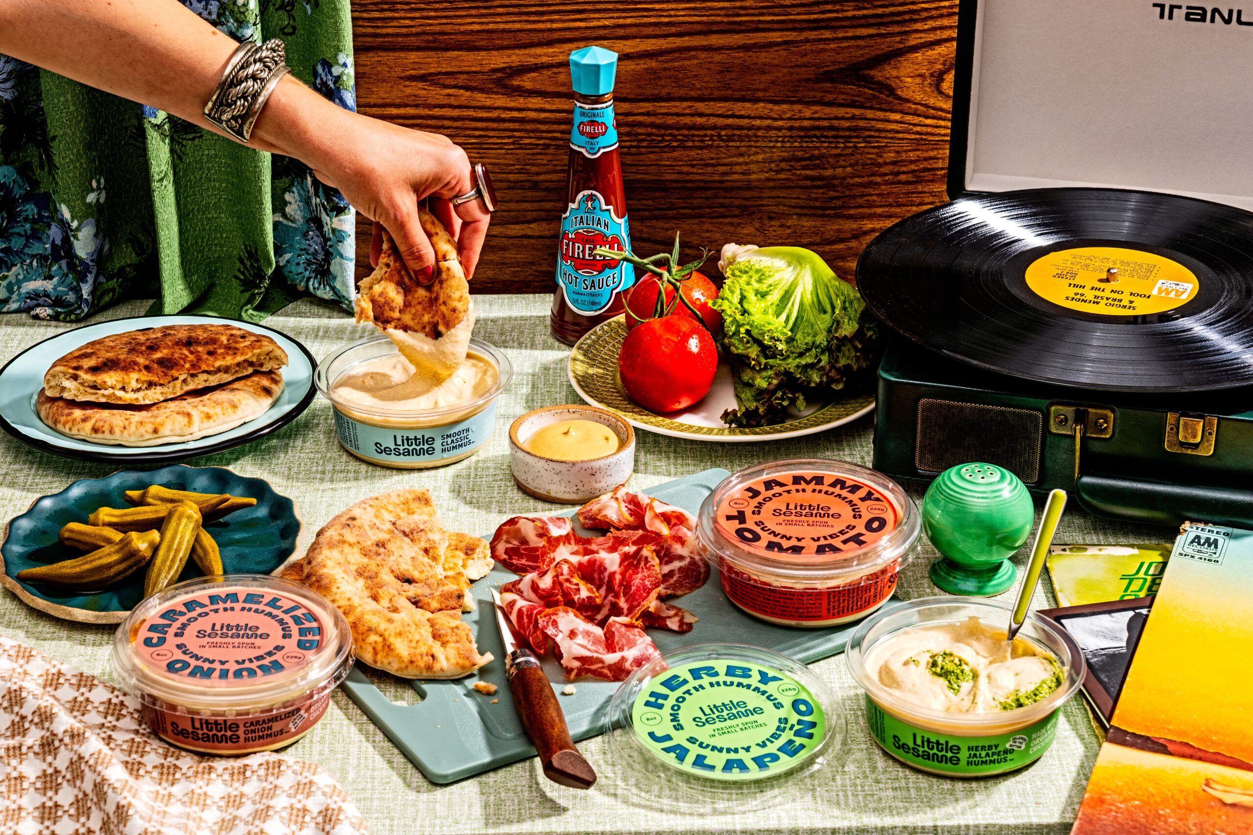 a model’s hand dips pita into one of little sesame hummus’, next to more of their spread alongside various foods and a record player.