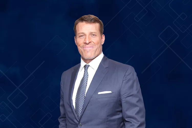 A man in a dark suit and tie stands against a dark blue geometric background, smiling at the camera.