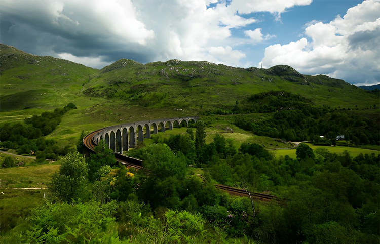 A long stone viaduct railway with arches stretches across a lush green landscape, set against rolling hills and a partly cloudy sky.