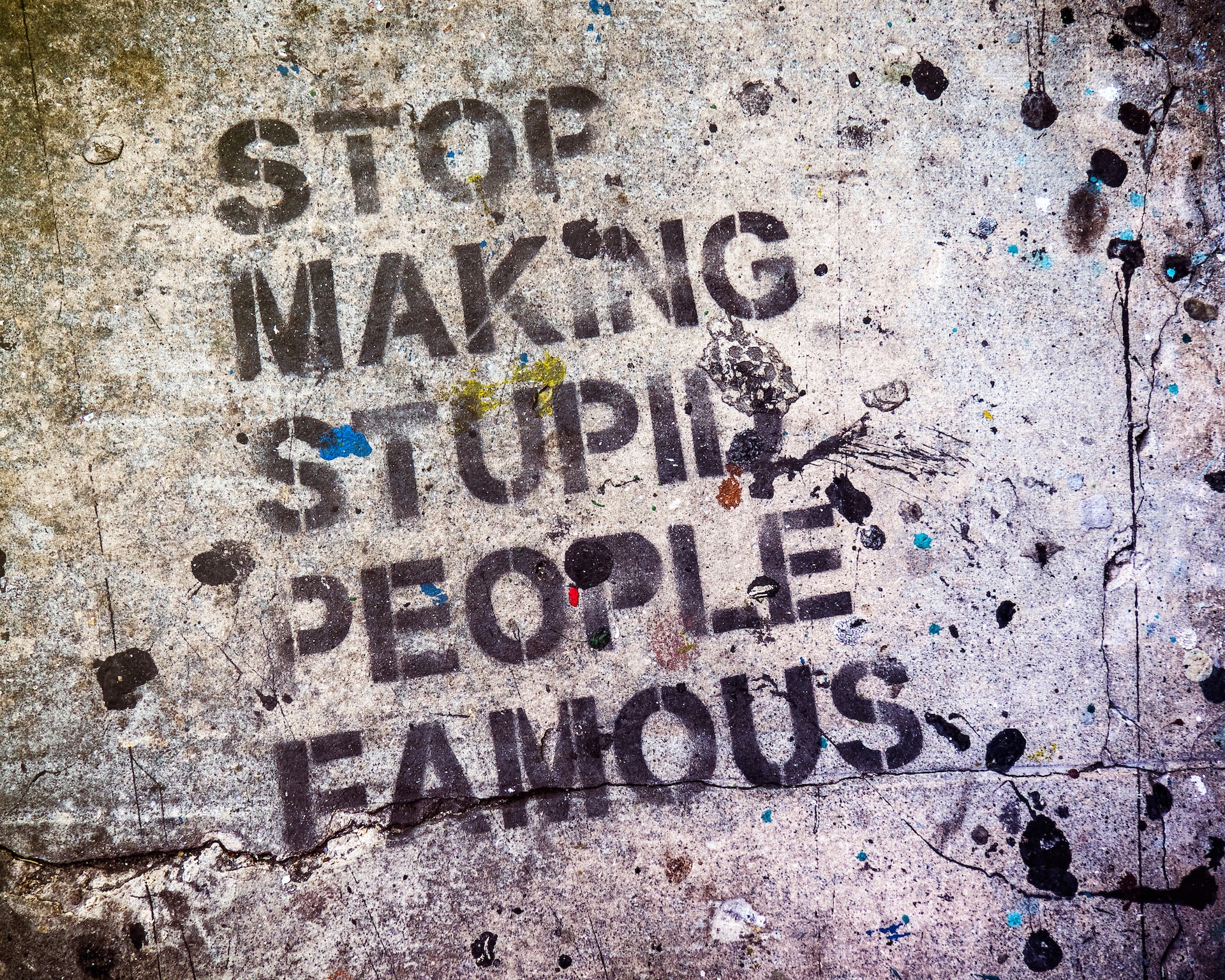 A stencil of the words “stop making stupid people famous” spray painted onto a concrete wall.