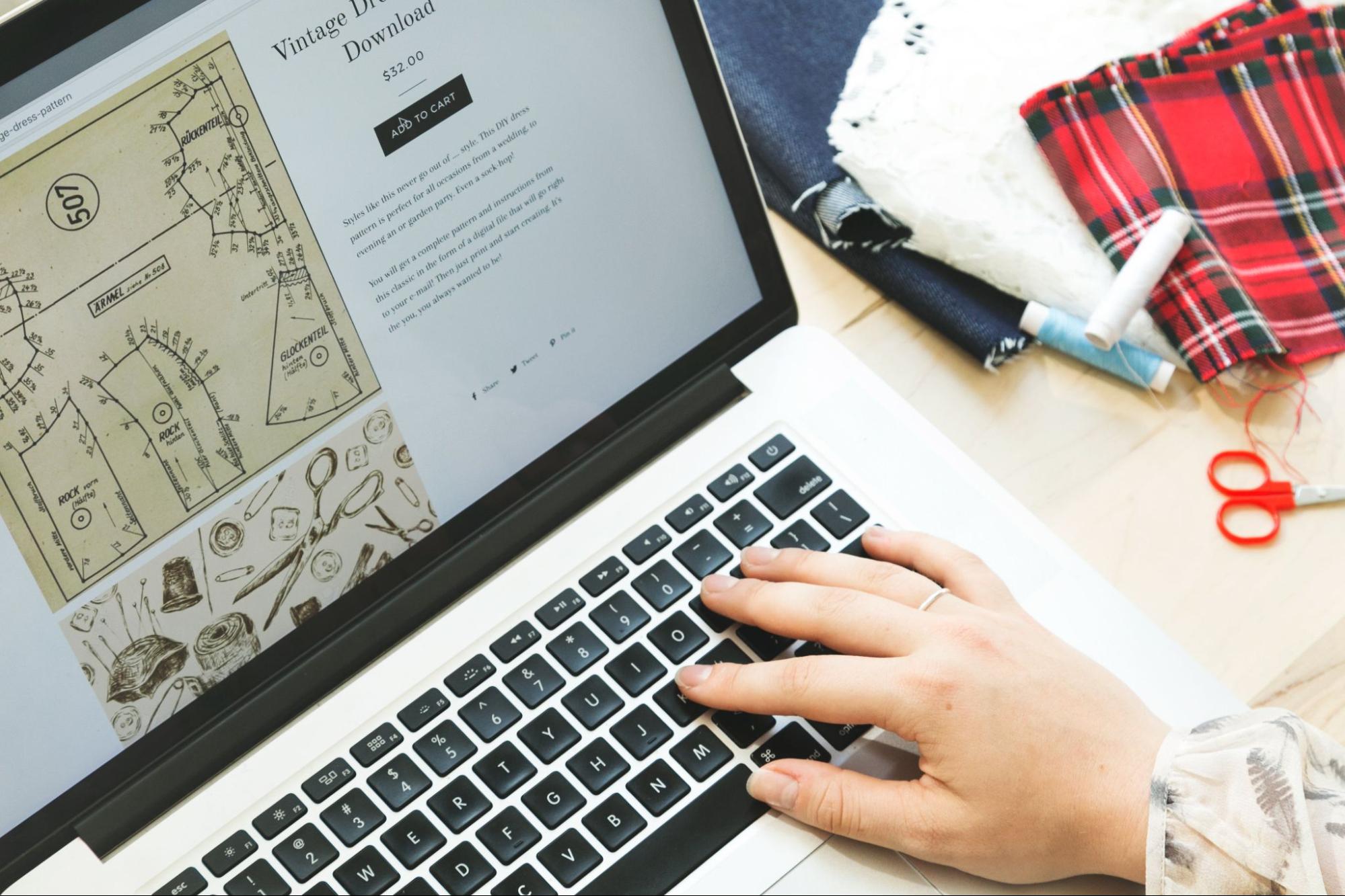 Next to a pile of fabrics, a hand reaches into the frame to order vintage dress patterns.