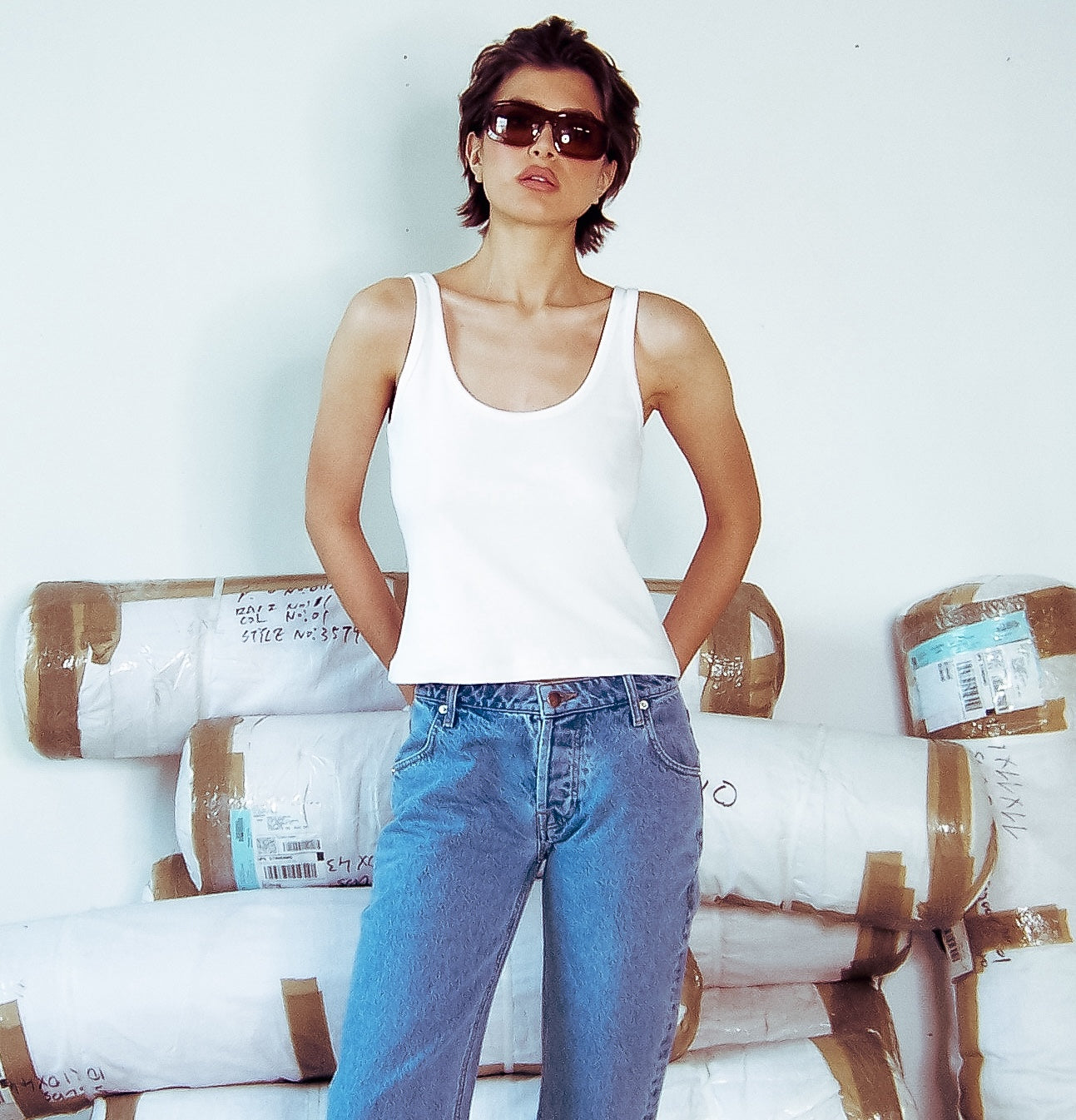A model poses wearing Uncle Studios white tank top and jeans in front of unopened packages.