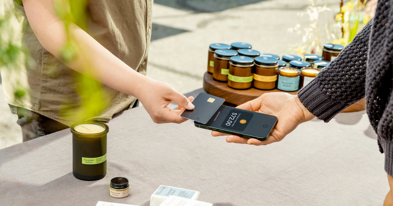 A customer taps a credit card on a smartphone to pay a vendor at an outdoor market.