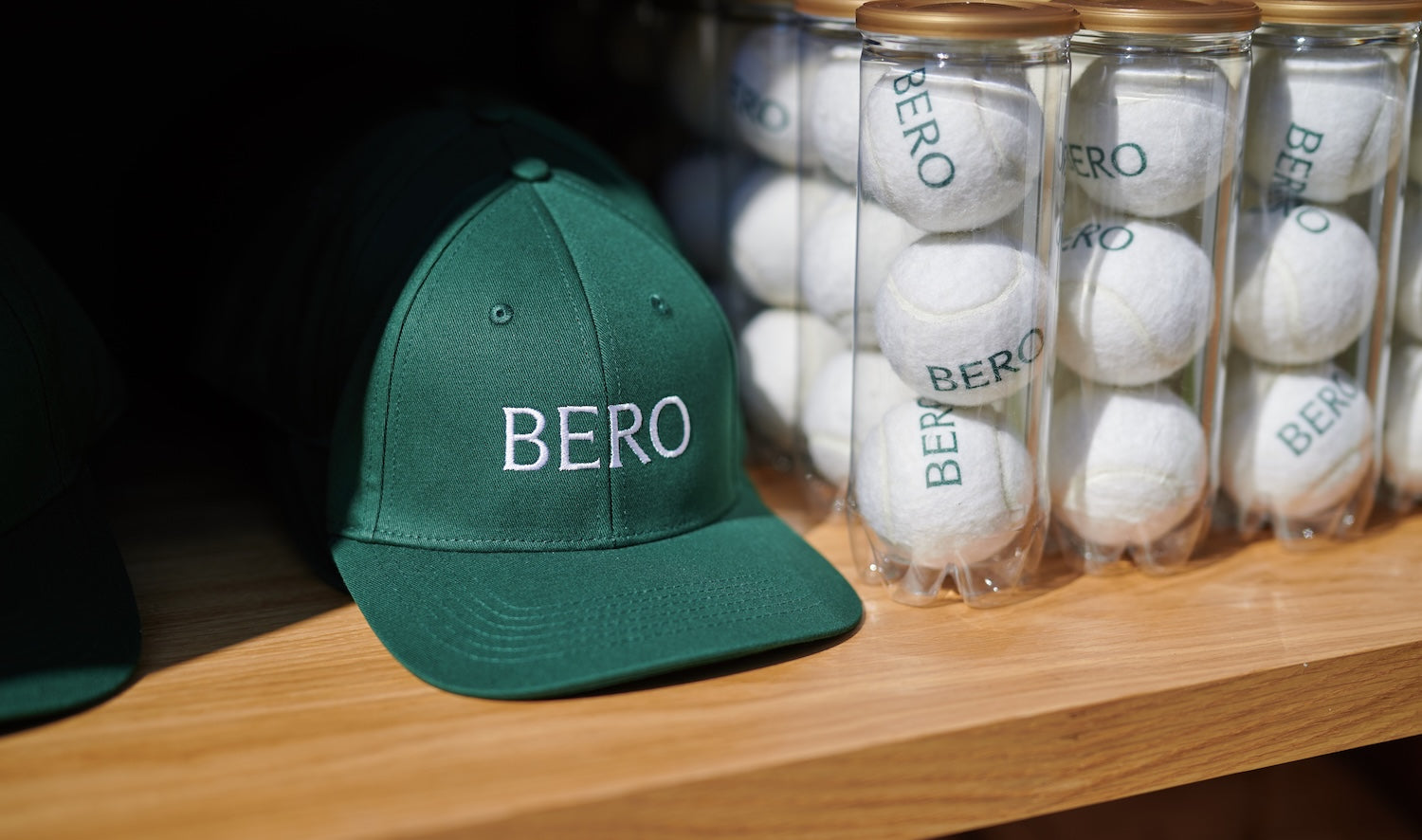 A green Bero branded hat sits on a shelf beside Bero branded tennsi balls